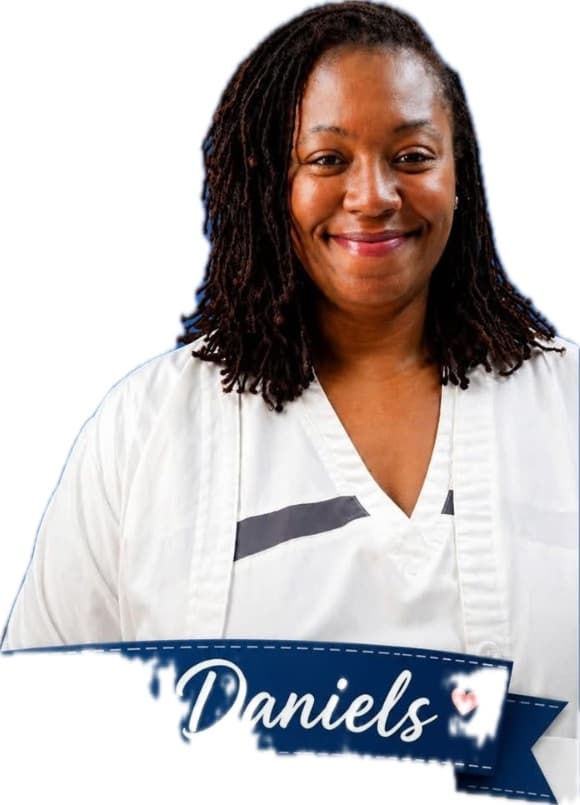 Smiling woman with long dark locs wearing white scrubs with a "Daniels" banner overlay.